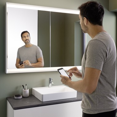 Un homme commande lʼéclairage de lʼarmoire à miroir Geberit ONE avec son smartphone (© Geberit)