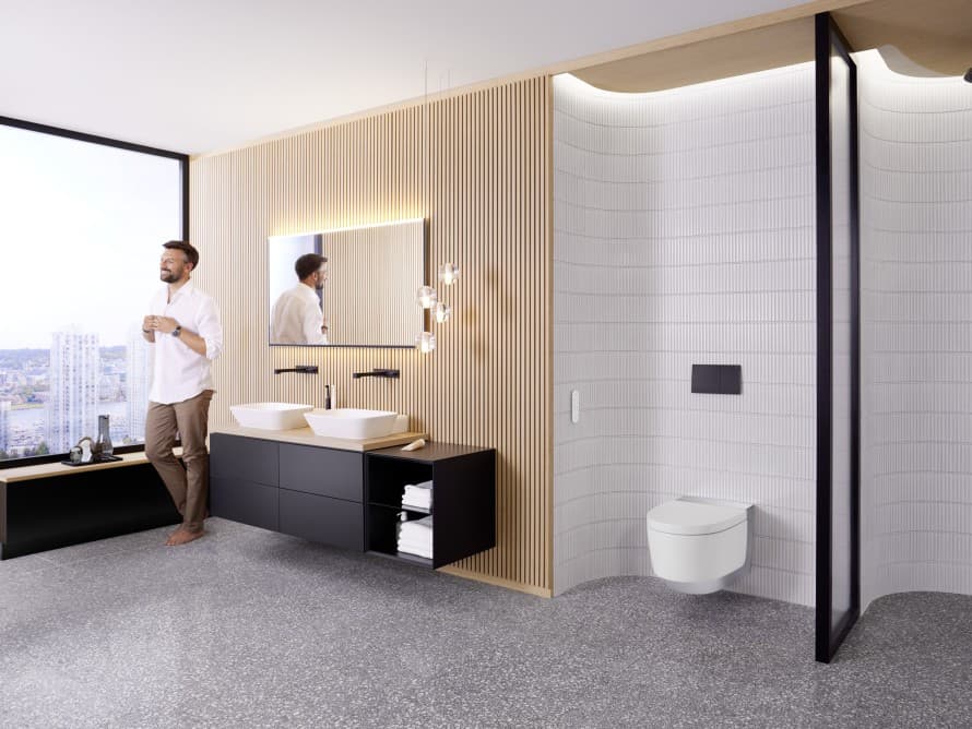 Homme dans une grande salle de bains avec meubles sombres et WC-douche Geberit AquaClean Homme dans une grande salle de bains avec meubles sombres et WC-douche Geberit AquaClean