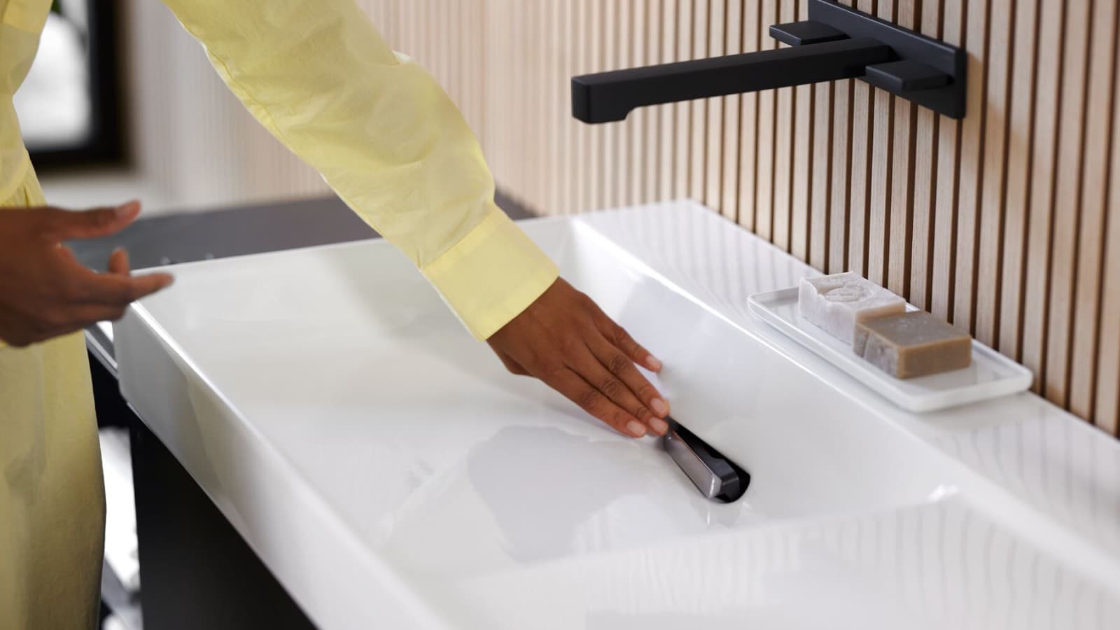 Une femme retire le filtre à cheveux sur un lavabo Geberit ONE avec robinet de lavabo en noir mat. (© Geberit) Une femme retire le filtre à cheveux sur un lavabo Geberit ONE avec robinet de lavabo en noir mat. (© Geberit)